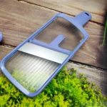 Mandolina cortadora de vegetales azul lavanda con cuchilla de acero expuesta, sobre fondo rústico de madera y hojas de eneldo.