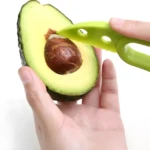 Manos usando cortador verde para remover el hueso de medio aguacate Hass cortado, fondo blanco.