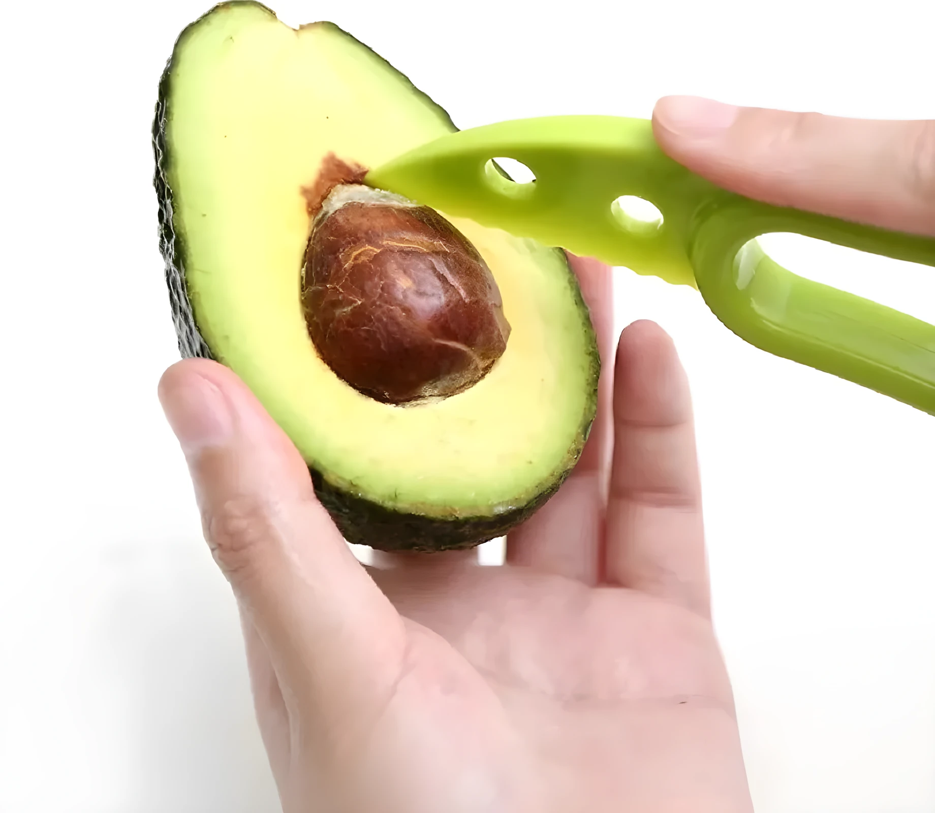 Manos usando cortador verde para remover el hueso de medio aguacate Hass cortado, fondo blanco.