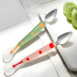 Dos utensilios doble cabeza bebé: cuchara silicona naranja y verde con puntos, y cuchara rosa y roja con borde dentado de metal.