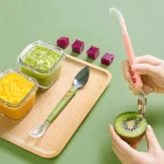 Set de dos utensilios para bebé: cuchara de silicona rosa y cuchillo/cuchara verde con sierra para kiwi, sobre bandeja de madera con purés de fruta.