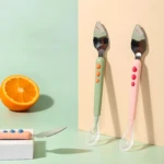 Juego de cucharas para bebé: una verde y una rosa con cabeza de metal serrada y punta de silicona, junto a una naranja cortada.