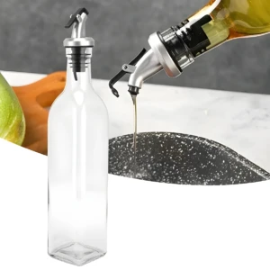 Botella dispensadora de aceite de vidrio vacía junto a otro envase vertiendo un chorro fino de aceite sobre una superficie oscura.