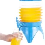 Dispensador de bebidas plegable azul y amarillo vertiendo jugo naranja en un vaso sostenido por una mano.