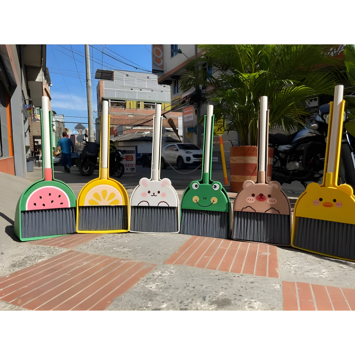 Escobas y recogedores de mano con diseño de frutas y animales (sandía, limón, oso, rana, pato) en colores vivos, sobre acera urbana.
