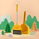 Escoba y recogedor amarillo infantil con diseño de pato sonriente, ambientado en un paisaje de papel recortado.