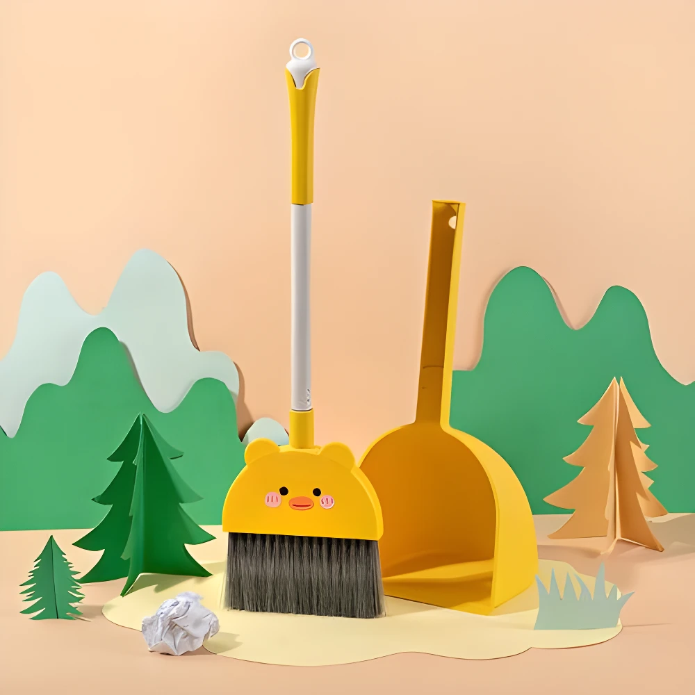 Escoba y recogedor amarillo infantil con diseño de pato sonriente, ambientado en un paisaje de papel recortado.