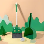 Set de escoba y recogedor infantil verde con diseño de rana sonriente, sobre fondo claro con decoración de naturaleza en papel.