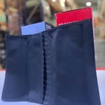 Faja reductora moldeadora negra con cierre frontal de ganchos ajustables, exhibida sobre una superficie blanca frente a cajas de producto en bodega.
