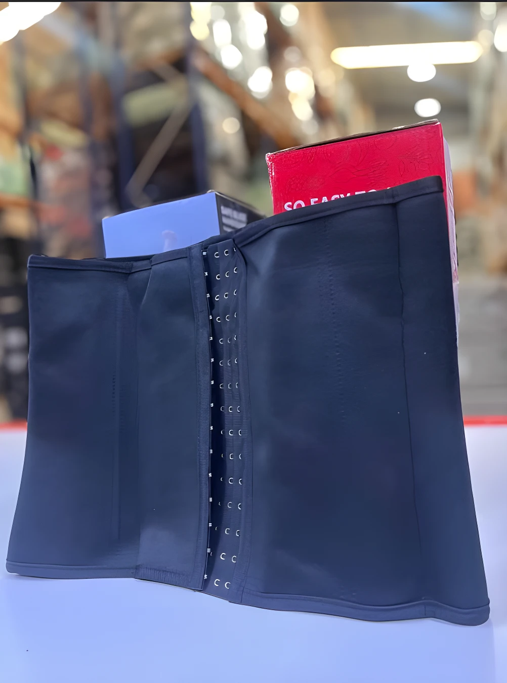 Faja reductora moldeadora negra con cierre frontal de ganchos ajustables, exhibida sobre una superficie blanca frente a cajas de producto en bodega.