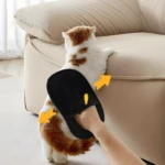 Mano usando guante negro de aseo para mascotas para cepillar el lomo de un gato atigrado naranja y blanco cerca de un sofá beige.