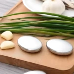 Tres jabones metálicos ovalados pulidos junto a hojas de cebollín y dientes de ajo sobre tabla de madera.