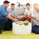 Familia jugando Spot It!, juego de cartas con latas verdes en mesa blanca sobre alfombra amarilla.