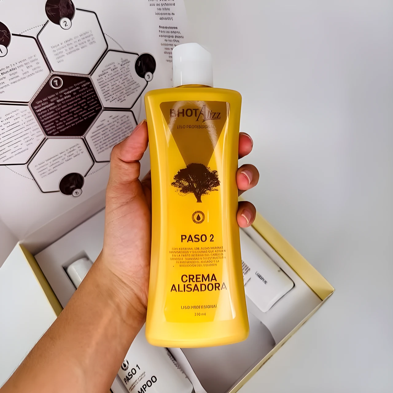 Botella amarilla de Crema Alisadora Bhot Alizz Professional con texto y árbol, sostenida por mano, parte de un kit.