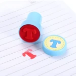 Sello de goma azul y rojo con la letra T estampada en rojo sobre hoja rayada junto a la tapa del sello.