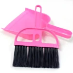 Kit escoba y recogedor plástico rosa brillante con cerdas negras, visto desde arriba sobre fondo blanco.