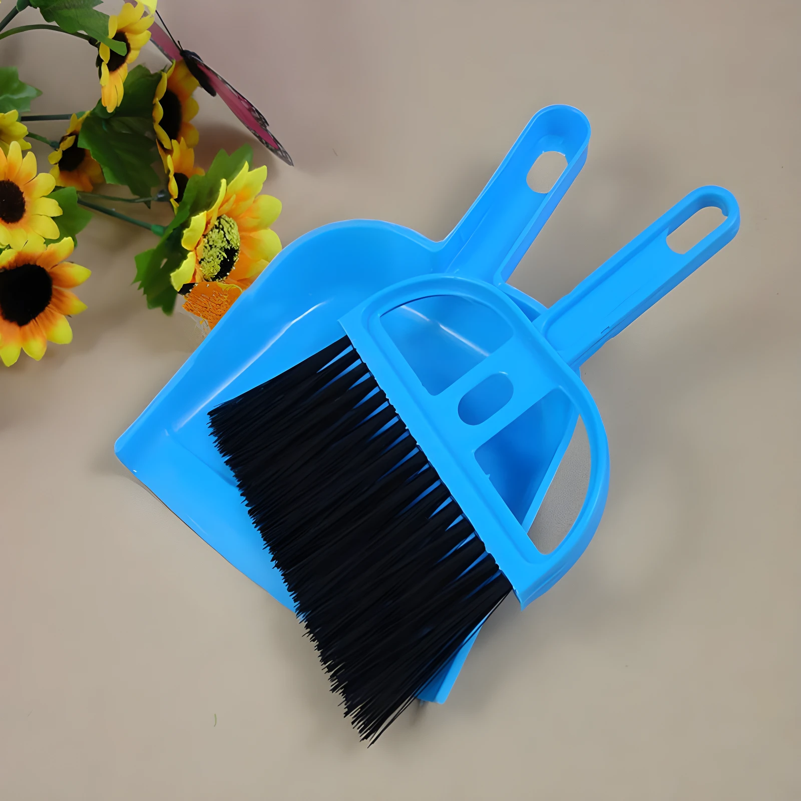 Kit escoba y recogedor pequeño azul brillante con cerdas negras, sobre fondo beige con girasoles artificiales.