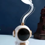 Lámpara de escritorio LED flexible con base figura de astronauta blanco y detalles dorados, luz blanca encendida, fondo azul oscuro.