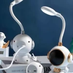 Dos lámparas LED de escritorio astronauta blancas y doradas. Una muestra el puerto de carga posterior. Fondo azul oscuro.