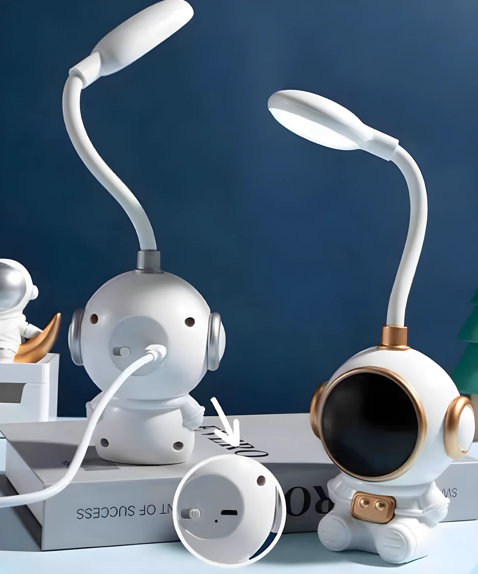 Dos lámparas LED de escritorio astronauta blancas y doradas. Una muestra el puerto de carga posterior. Fondo azul oscuro.