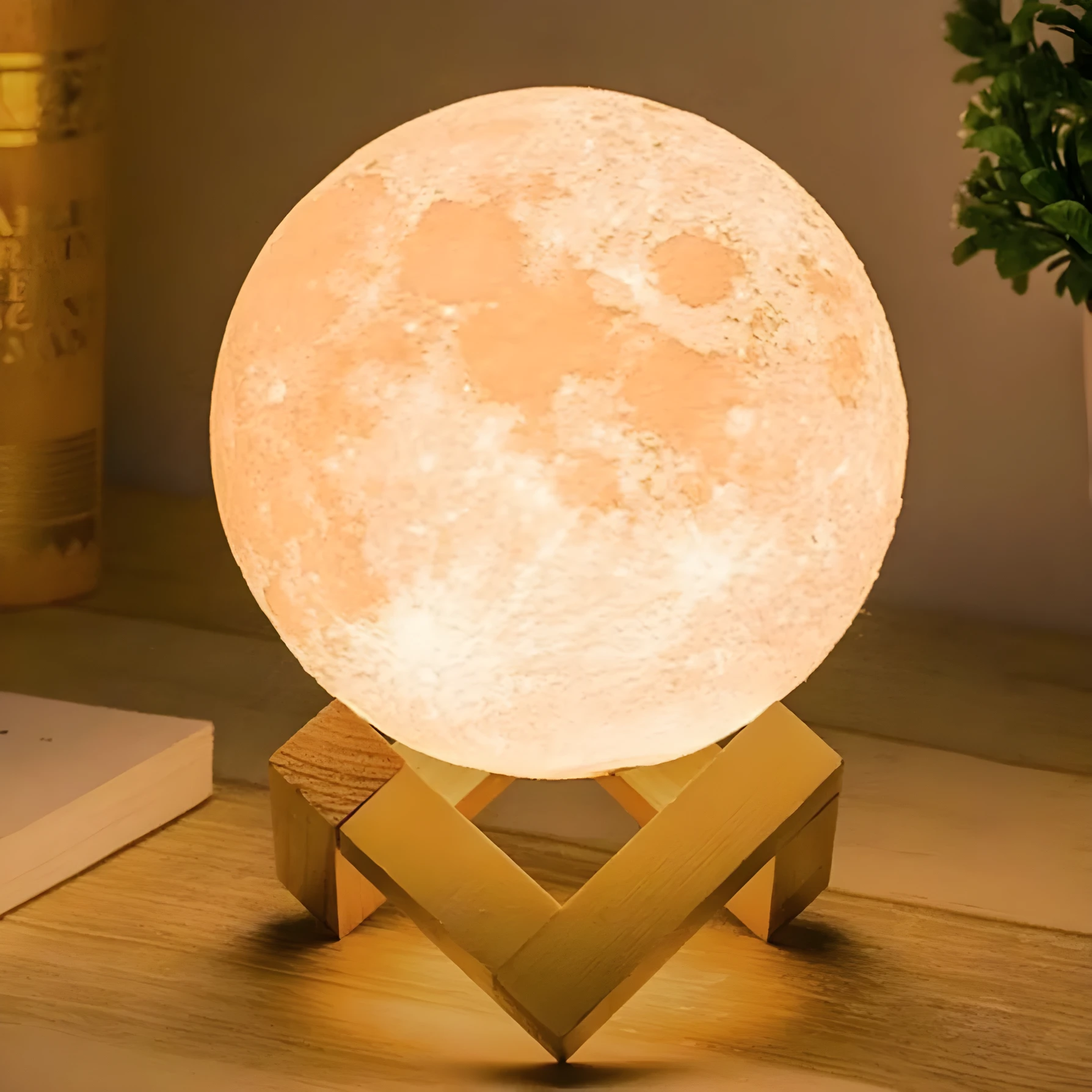 Lámpara de luna 3D encendida, tono naranja cálido, sobre base de madera clara en mesa junto a libro y planta.