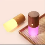 Lámpara de mesa cilíndrica con base de madera oscura y luz inferior magenta, junto a otra con luz amarilla tenue.