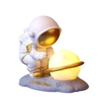 Lámpara noche astronauta blanco y dorado, inclinado junto a esfera luminosa tipo planeta con anillo. Base lunar con texto "Little Star".