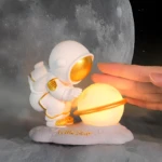 Lámpara noche astronauta blanco con detalles dorados cuidando una esfera luminosa tipo planeta con anillo.