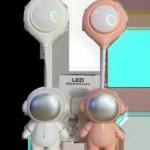 Dos lámparas LED de escritorio con figura de astronauta, una blanca y otra rosa, con brazo flexible y luz superior circular, junto a su caja.