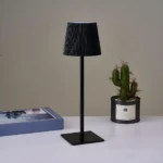 Lámpara LED negra minimalista de metal, inalámbrica, con pantalla acanalada, sobre mesa blanca junto a cactus.
