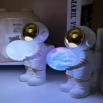Dos lámparas astronauta sostienen esferas luminosas, una blanca y otra colorida, en un estante con libros y decoración.