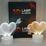 Lámparas 3D de corazón flechado, una blanca y otra amarilla, sobre bases circulares blancas, frente a cajas de regalo.