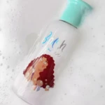 Líquido limpiador de brochas en botella transparente con personaje y tapa turquesa, rodeado de espuma.