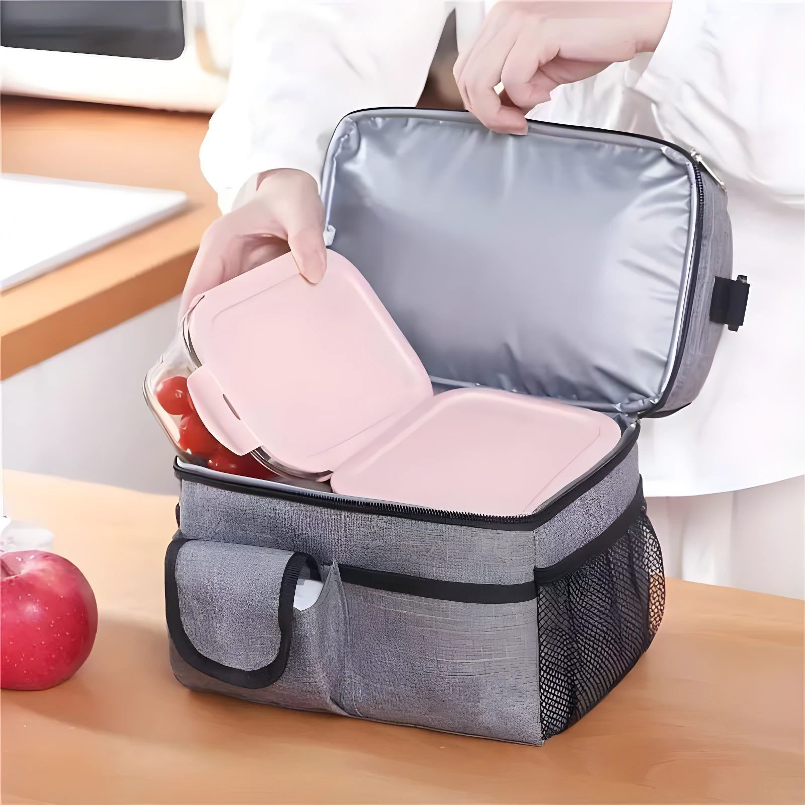 Lonchera térmica gris con compartimentos aislados, persona colocando recipiente rosa con tomates.