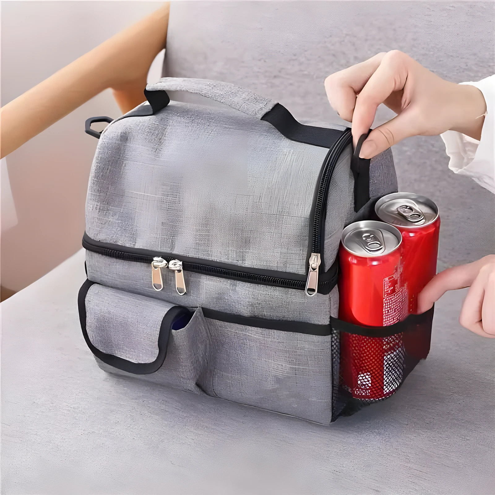 Lonchera térmica gris de doble compartimiento, con dos latas de refresco en el bolsillo lateral de malla.