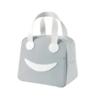Lonchera térmica portátil gris de diseño cuadrado con carita feliz, dos asas blancas superiores y cierre lateral, ideal para transportar alimentos.