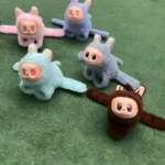 Cuatro peluches coleccionables estilo muñeco con orejas, rostro de vinilo y cuerpo de felpa en colores rosa, azul claro, azul grisáceo y marrón, sobre césped artificial verde.