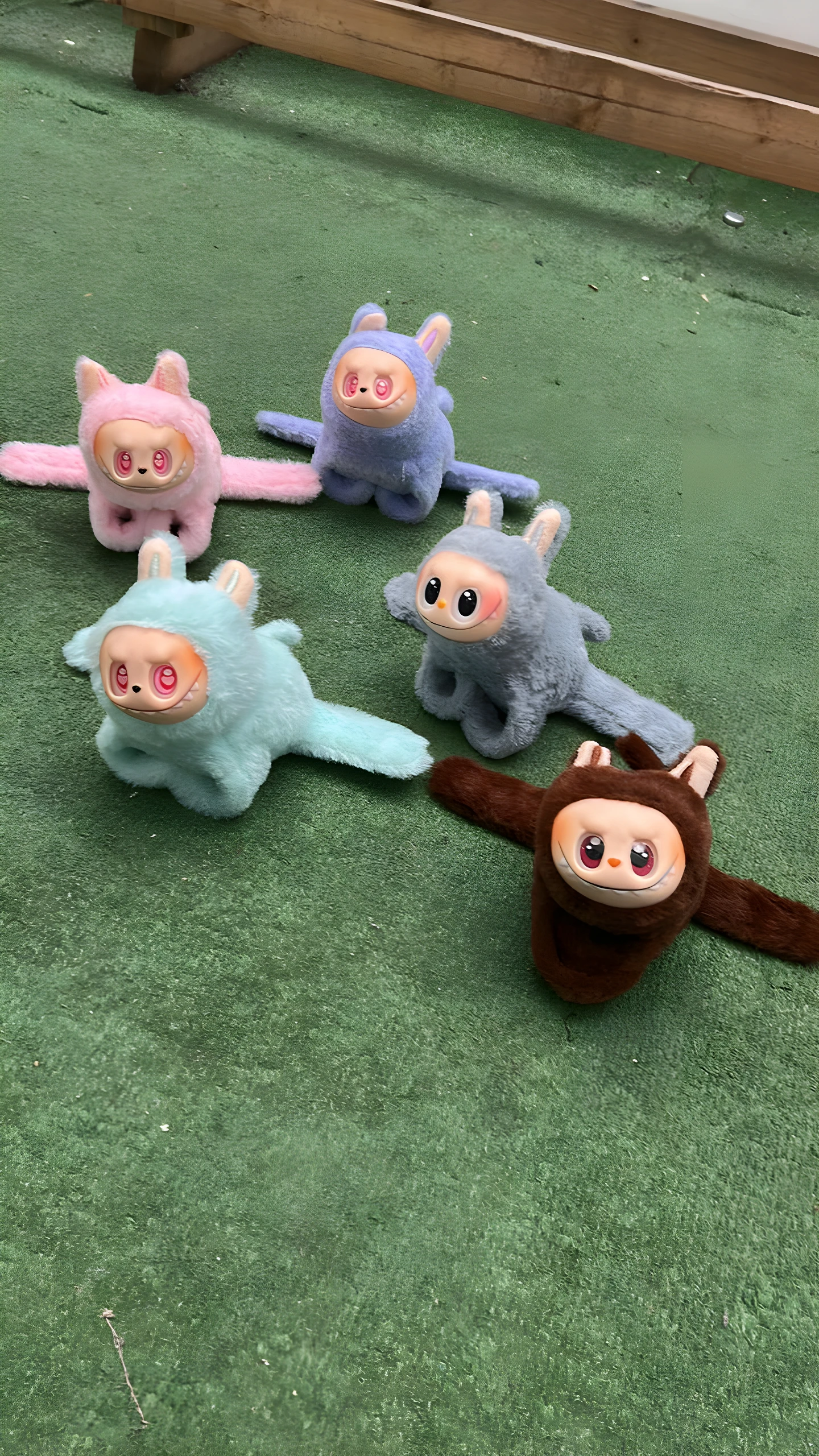 Cuatro peluches coleccionables estilo muñeco con orejas, rostro de vinilo y cuerpo de felpa en colores rosa, azul claro, azul grisáceo y marrón, sobre césped artificial verde.