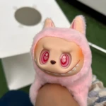 Peluche coleccionable Labubu rosa con capucha peluda, ojos rojos brillantes con corazón, sobre pierna humana, con caja blanca al fondo.