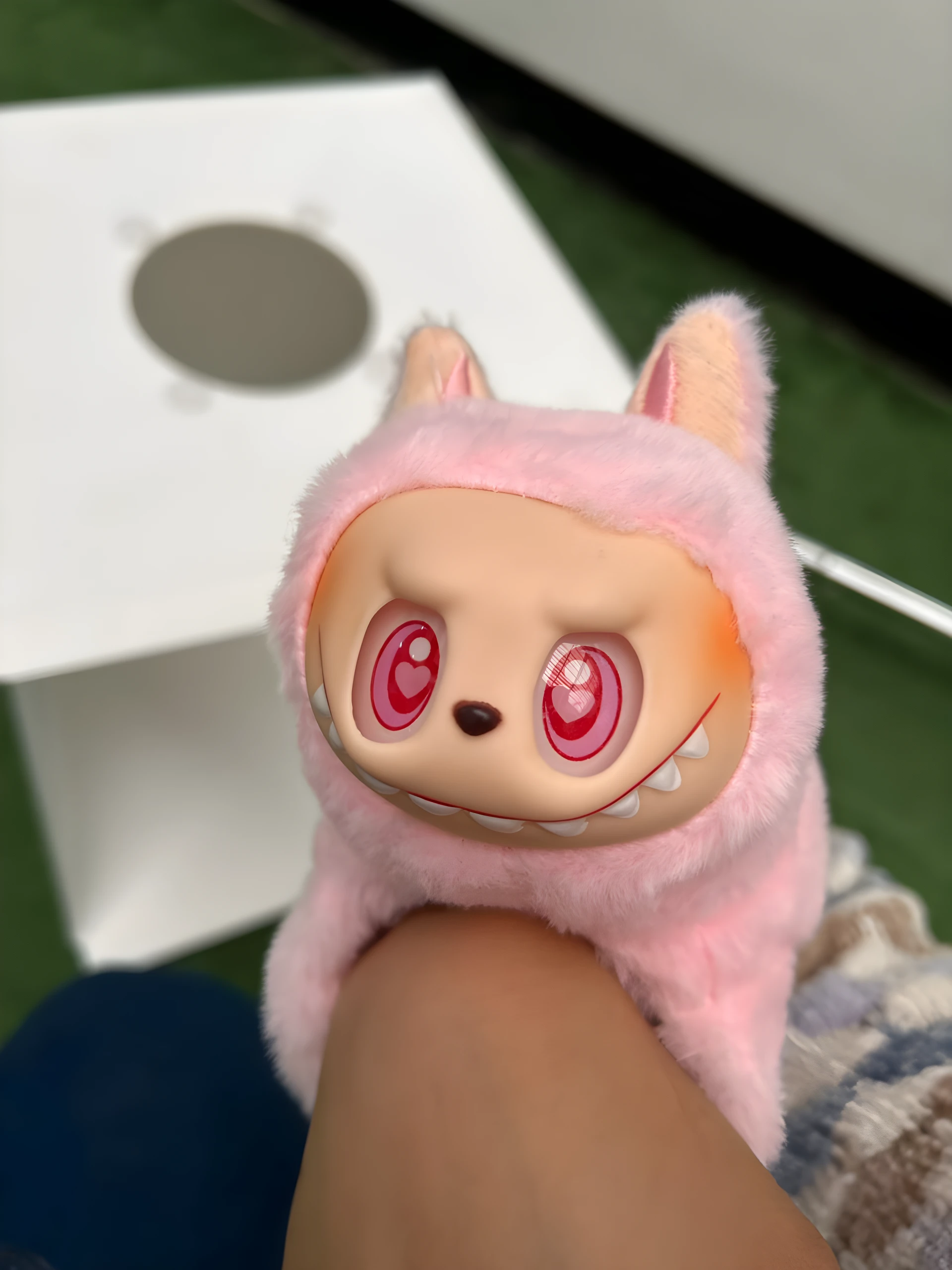 Peluche coleccionable Labubu rosa con capucha peluda, ojos rojos brillantes con corazón, sobre pierna humana, con caja blanca al fondo.