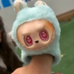 Manilla de peluche suave azul menta con cabeza de personaje Labubu de ojos rojos en forma de corazón, siendo sostenida en una mano.
