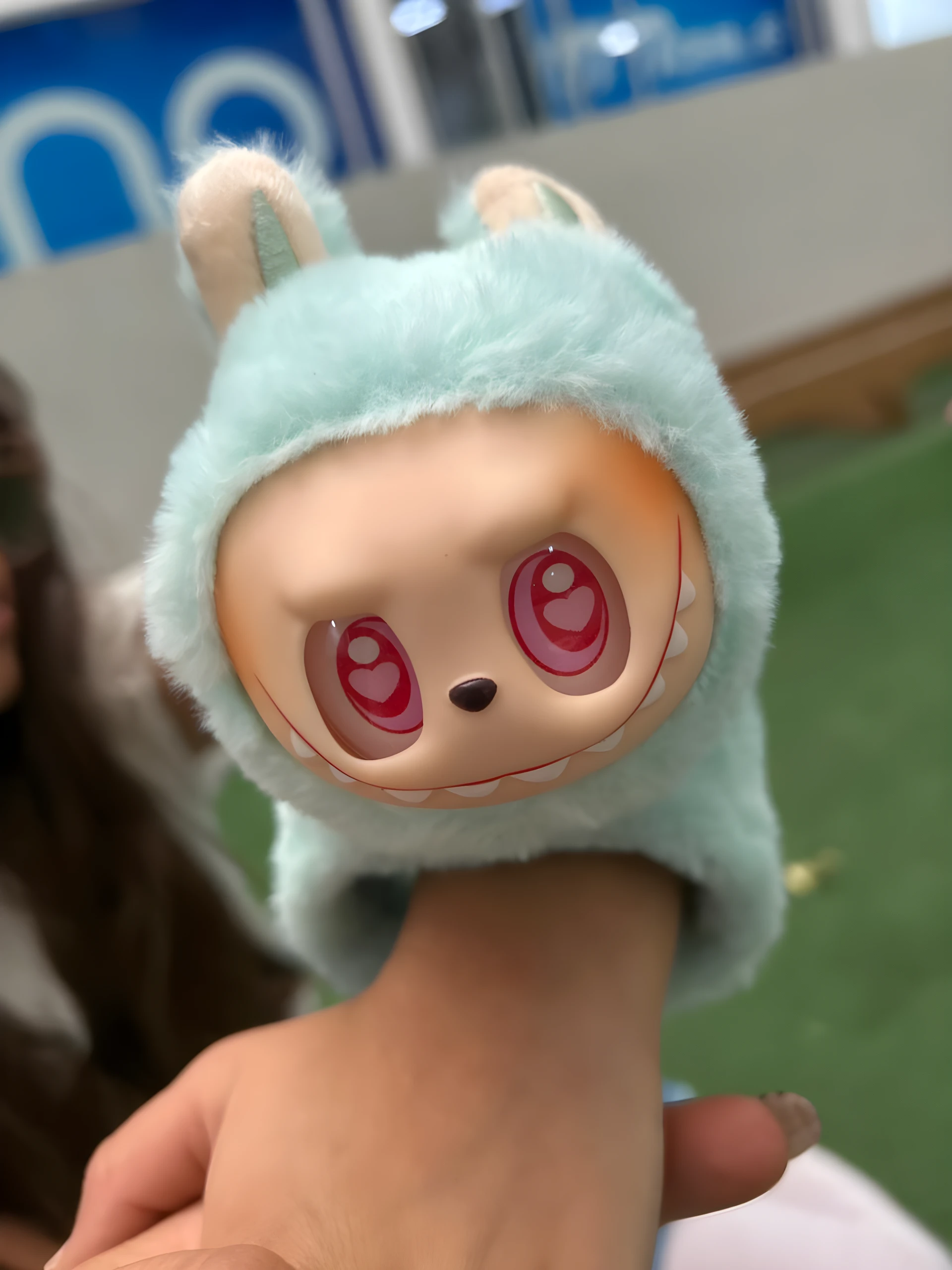 Manilla de peluche suave azul menta con cabeza de personaje Labubu de ojos rojos en forma de corazón, siendo sostenida en una mano.