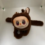 Peluche Labubu marrón peludo con orejas, rostro de vinilo blanco, ojos grandes morados brillantes y sonrisa con dientes puntiagudos, colgando sobre fondo blanco.