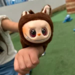 Manilla peluche marrón oscuro con cabeza de personaje sonriente, dientes blancos y ojos grandes rojos, sostenida por una mano sobre fondo verde.