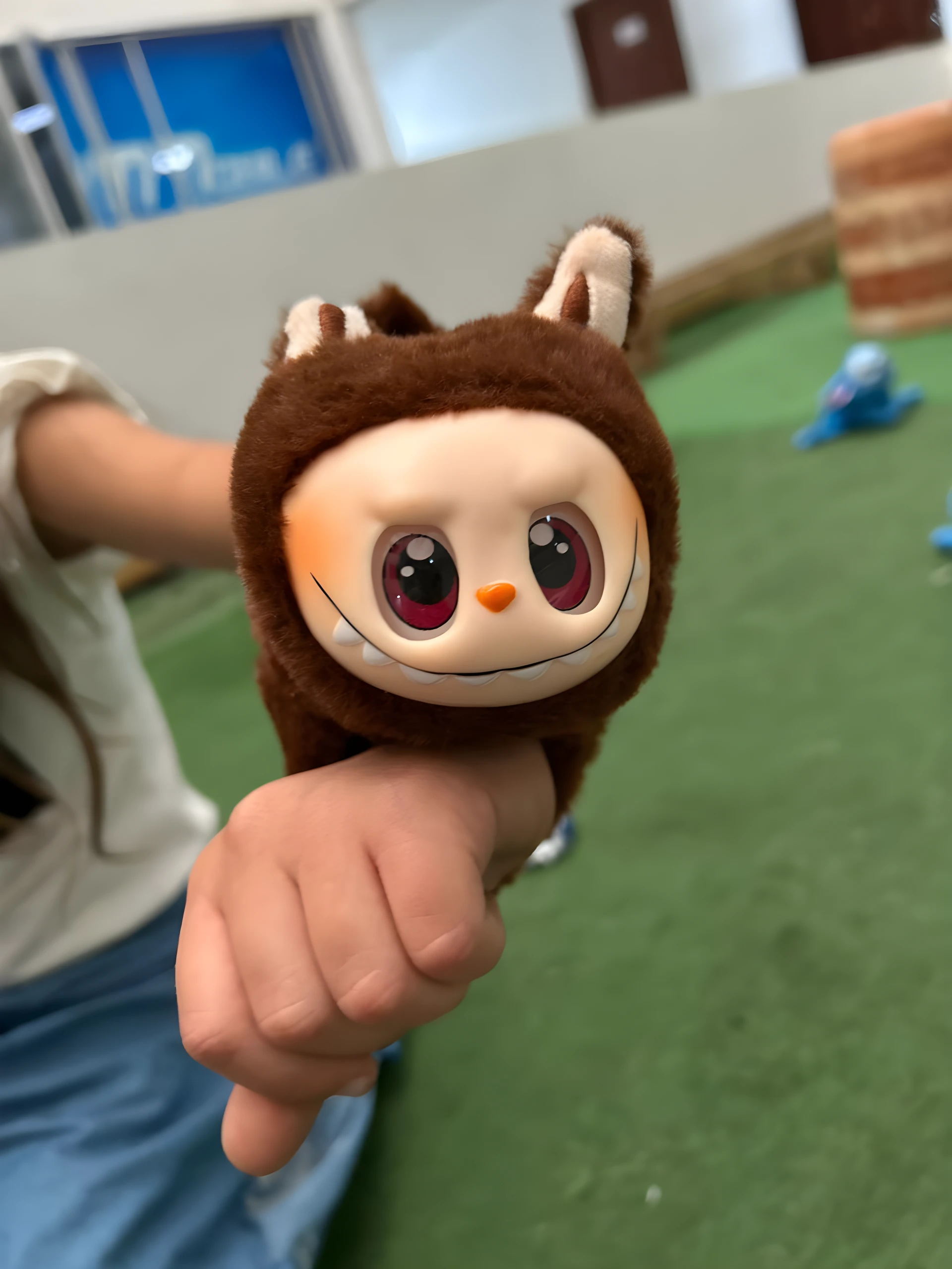 Manilla peluche marrón oscuro con cabeza de personaje sonriente, dientes blancos y ojos grandes rojos, sostenida por una mano sobre fondo verde.