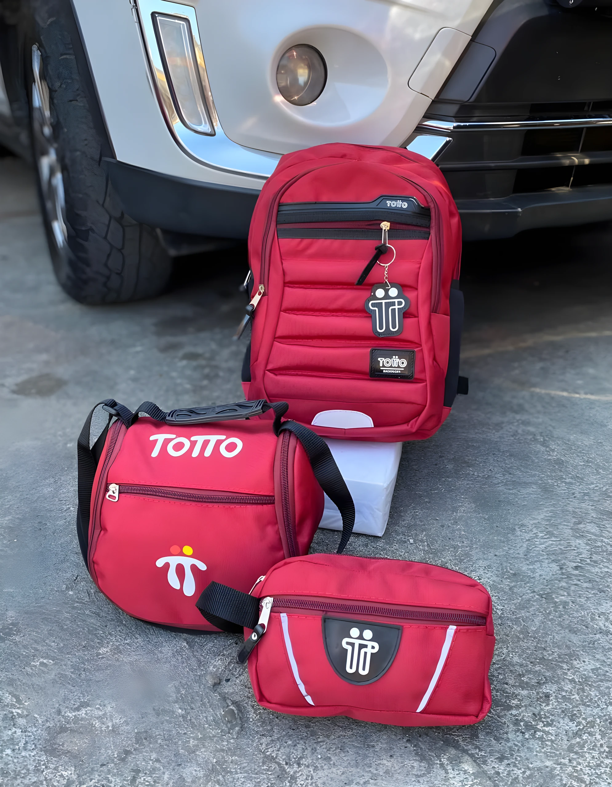 Morral Totto rojo acolchado con llavero temático, lonchera y cartuchera a juego, al frente de un vehículo.