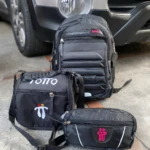 Tres morrales Totto negros: una cartuchera, una lonchera y un morral grande, dispuestos sobre un concreto al lado de un auto.