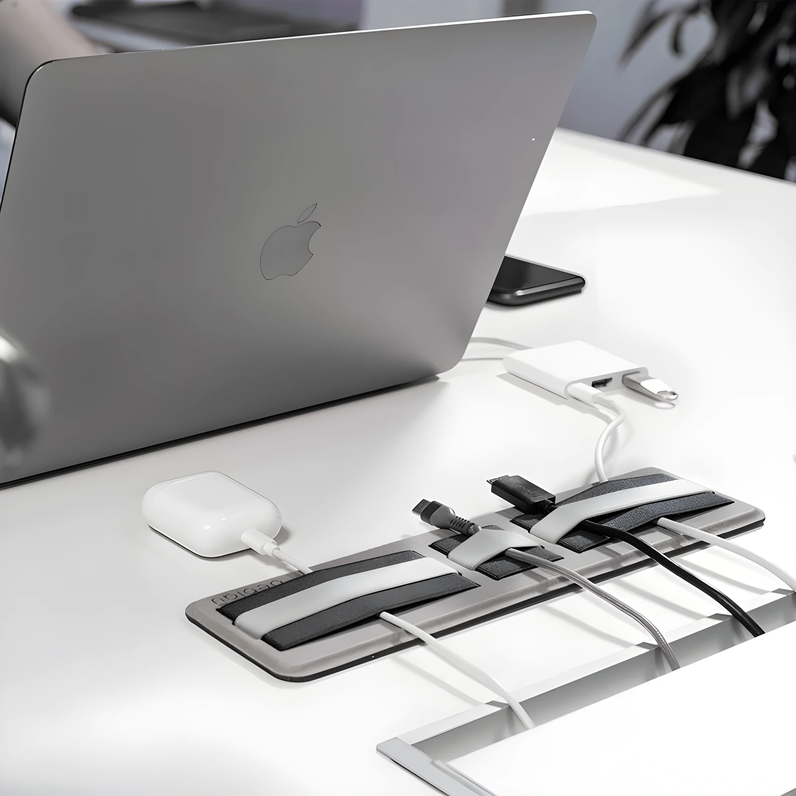 Organizador adhesivo para cables y accesorios junto a laptop Apple plateada, AirPods y adaptadores en escritorio blanco.
