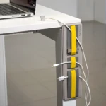 Organizador adhesivo gris con detalles amarillos para cables, sujetando cargadores de celular y laptop en un escritorio.