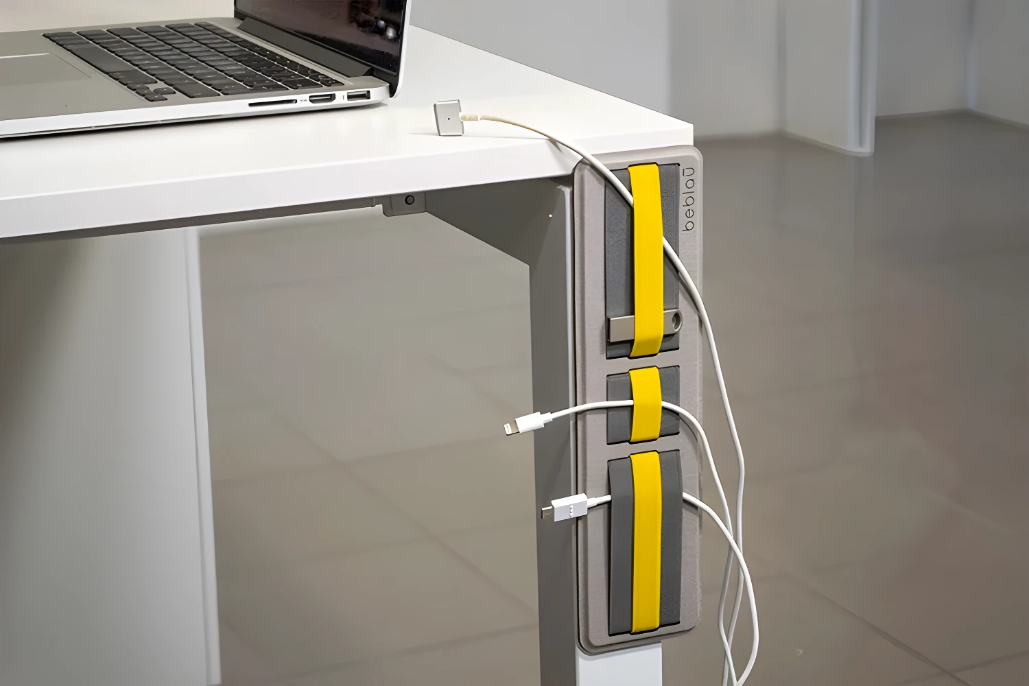 Organizador adhesivo gris con detalles amarillos para cables, sujetando cargadores de celular y laptop en un escritorio.